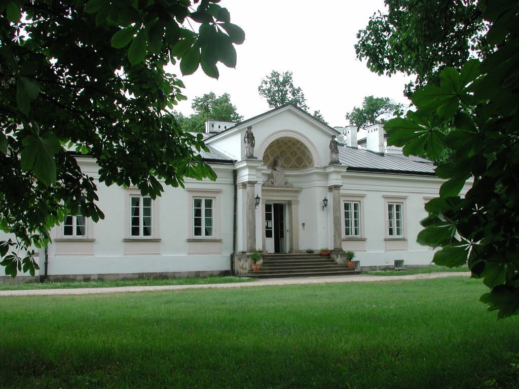 Zdjęcie przedstawia front białego pałacu. Na pierwszym planie jest zielony trawnik i gałęzie kasztanowca.