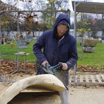 Artysta przy pracy na placu rzeźbiarskim. Używa piły spalinowej pracując nad dużym kawałkiem drewna. Ujęcie z innego kąta.