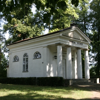 Jest lato. Zdjęcie przedstawia kaplicę w parku. Jest biała i ma 4 kolumny od frontu.