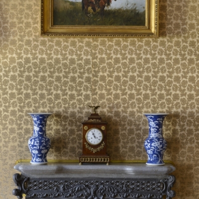 Pałac. Ściana w jadalni. Góra  kominka z półką. Na półce 2 niebieskie wazony, pomiędzy nimi zegar. Nad nimi wisi niewielki obraz – jeździec na koniu w stepie. 