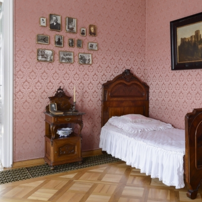 Pałac. Wnętrze dziewiętanstowiecznej sypialni. Jest tu zaścielone łóżko, nad nim obraz, obok głowy łóżka na ścianie 13 oprawionych fotografii rodzinnych. 